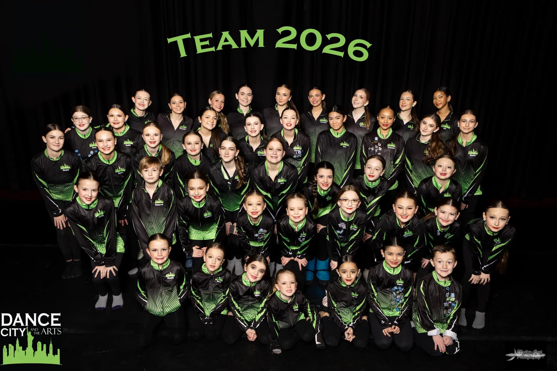 Dance City & the Arts Competitive Dance Team 2026 — full team in black and green warm-up jackets posed on stage in front of a TEAM 2026 backdrop.